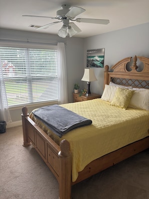 A bedroom with a bed, a ceiling fan, a window with blinds, a nightstand with a lamp, and a painting on the wall.