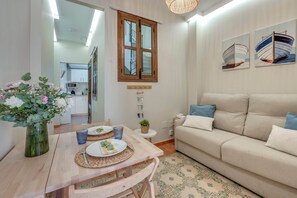 A living room with a sofa, a dining table with plates and cutlery, a vase with flowers, and a window with a view of the outside. - Cozy and lovely apartment located in the historic center of Málaga. (Málaga)