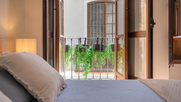 A room with a bed, a balcony with hanging plants, and a view of a building with arched windows. - Piso N°3 - Oliva Apartments Molinillo (Centro Histórico)