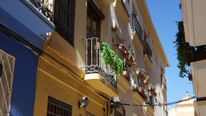 A narrow street with buildings featuring balconies with potted plants, a blue and yellow facade, and a tiled pavement. - Piso N°3 - Oliva Apartments Molinillo (Centro Histórico)