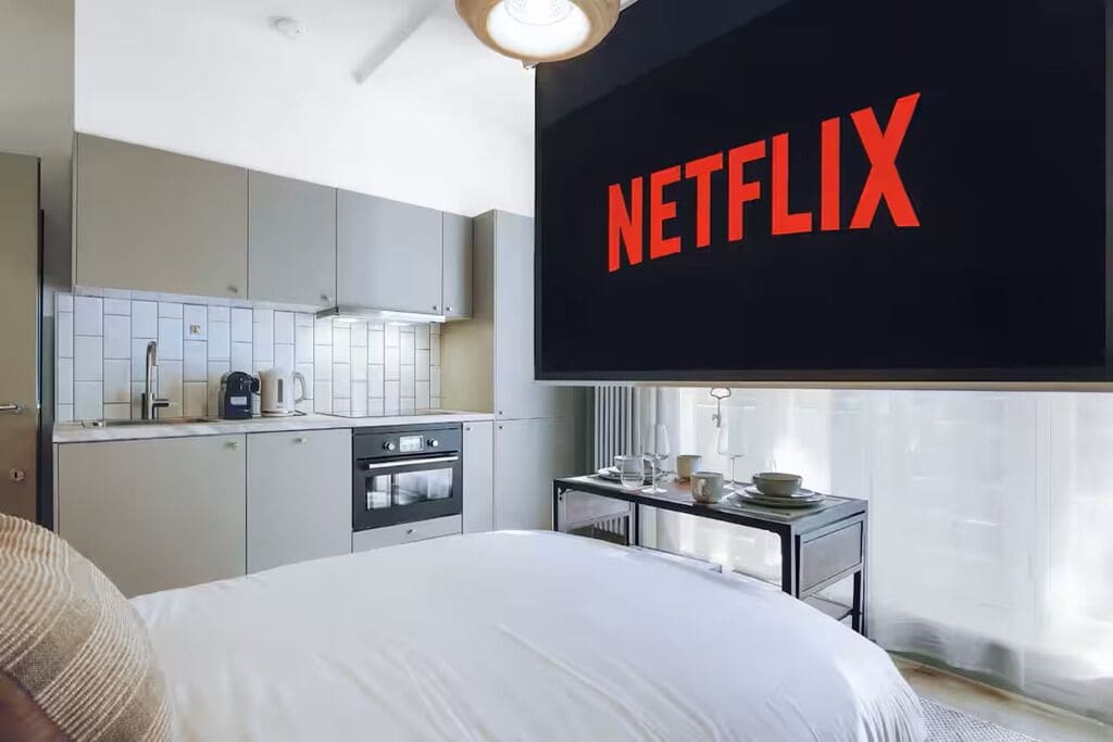 A hotel room with a bed, a kitchenette, and a large screen displaying the Netflix logo.