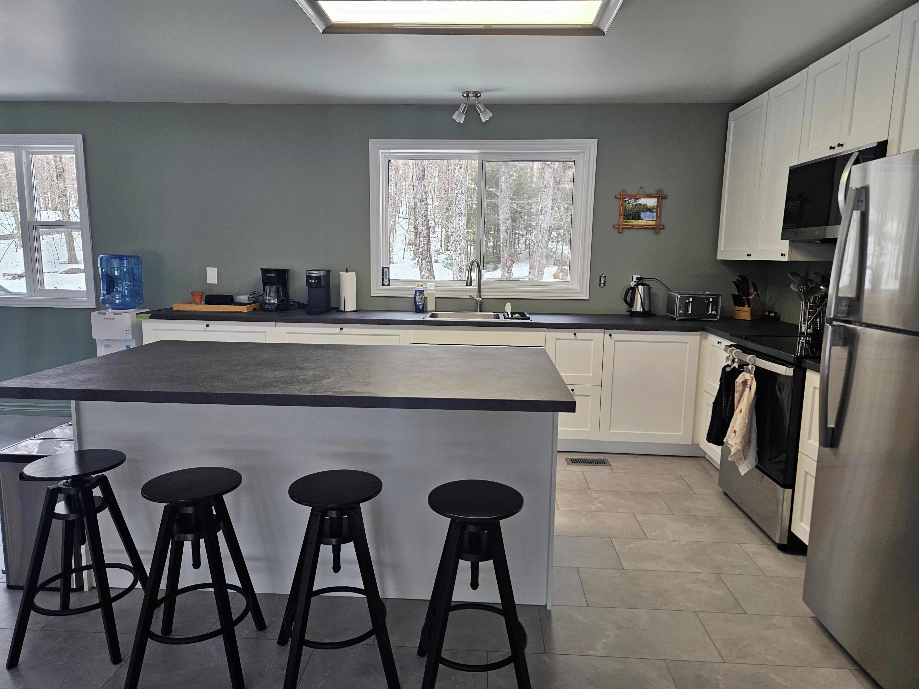 A modern kitchen with a large island, bar stools, a refrigerator, and a window with a view of trees.