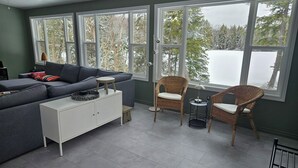 Uma sala de estar moderna com um sofá cinza, um armário branco, cadeiras de vime e grandes janelas com vista para árvores cobertas de neve.