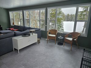 Ein modernes Wohnzimmer mit einem grauen Sofa, einem weißen Schrank, Korbsesseln und großen Fenstern mit Blick auf schneebedeckte Bäume.