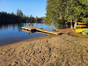 A sandy beach with a wooden dock extending into a calm lake, surrounded by trees. - Family friendly lakeside cottage. (Kearney)