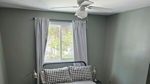 Um quarto com cama, mesa de cabeceira, luminária, janela com cortinas e ventilador de teto.