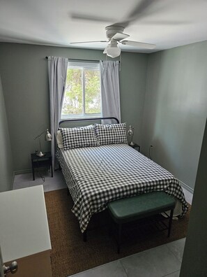 Ein Schlafzimmer mit einem Bett, einem Nachttisch, einer Lampe, einem Fenster mit Vorhängen und einem Deckenventilator.