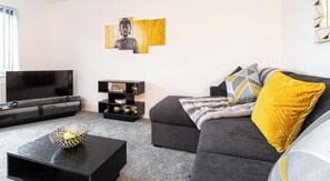 A living room with a flat-screen TV, a black entertainment unit, a grey sofa with patterned cushions, and a yellow throw pillow. - 6 Tannery House | Wi-Fi | Parking | Contractors (Freckleton)