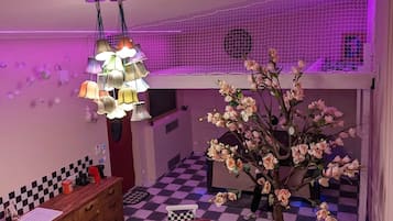 Huoneessa on ruutukuviollinen lattia, puinen kaappi, ruutukuviollisella pöytäliinalla katettu pöytä, tuoli, koristeellinen puu ja katosta roikkuva valaisin.