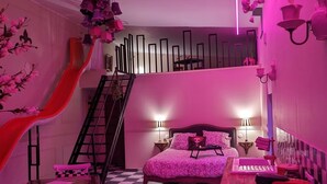 Een kamer met een zwart-wit geruite vloer, een bed met een roze sprei, een bureau met een computer, een stoel, een kleine tafel, een trap en een wandplank met decoratieve objecten.