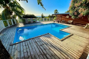 A wooden deck with a swimming pool, surrounded by a white fence and palm trees.