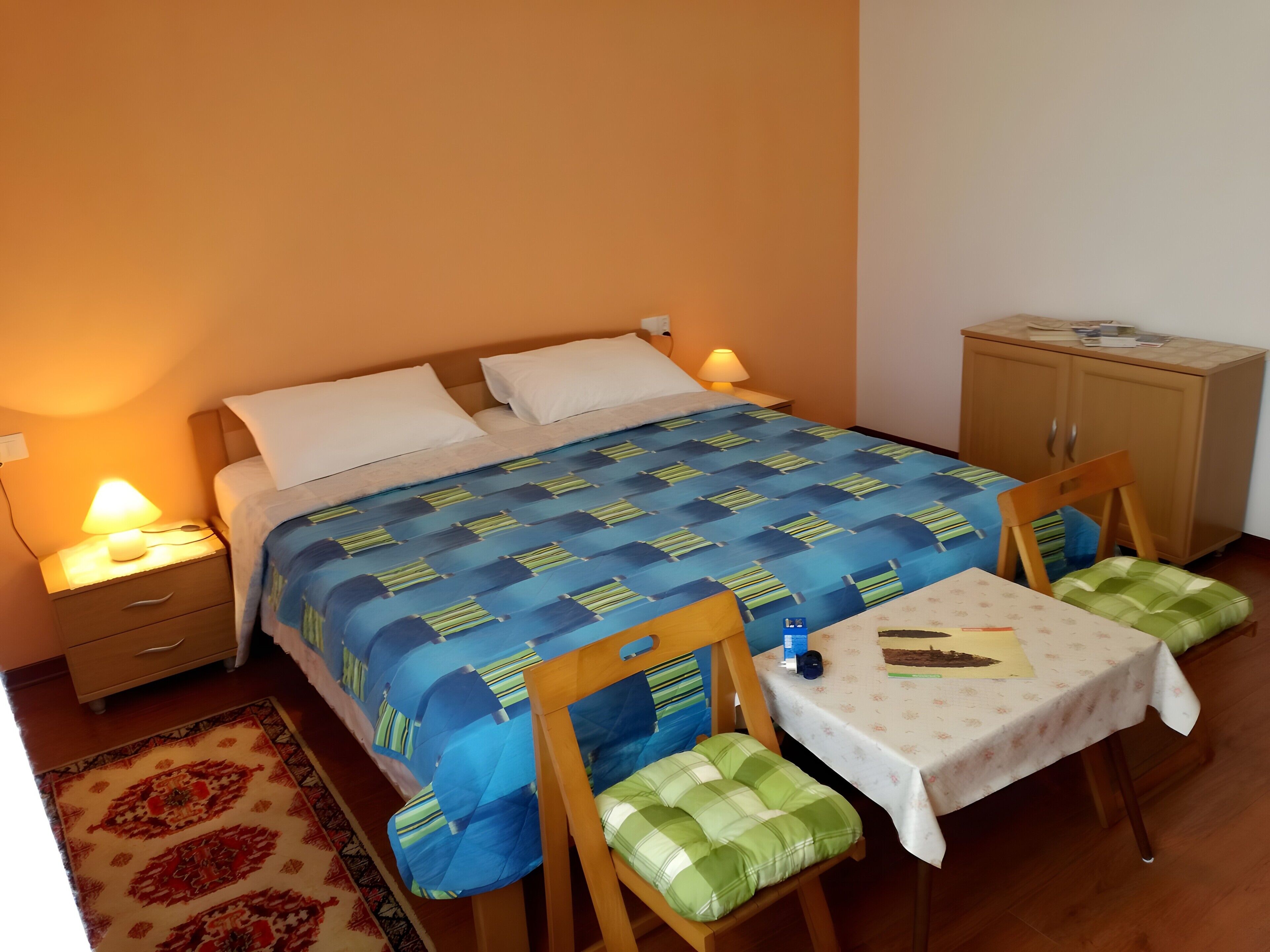 A bedroom with a bed, bedside tables, a nightstand, a wooden cabinet, a dining table with chairs, and a patterned rug.