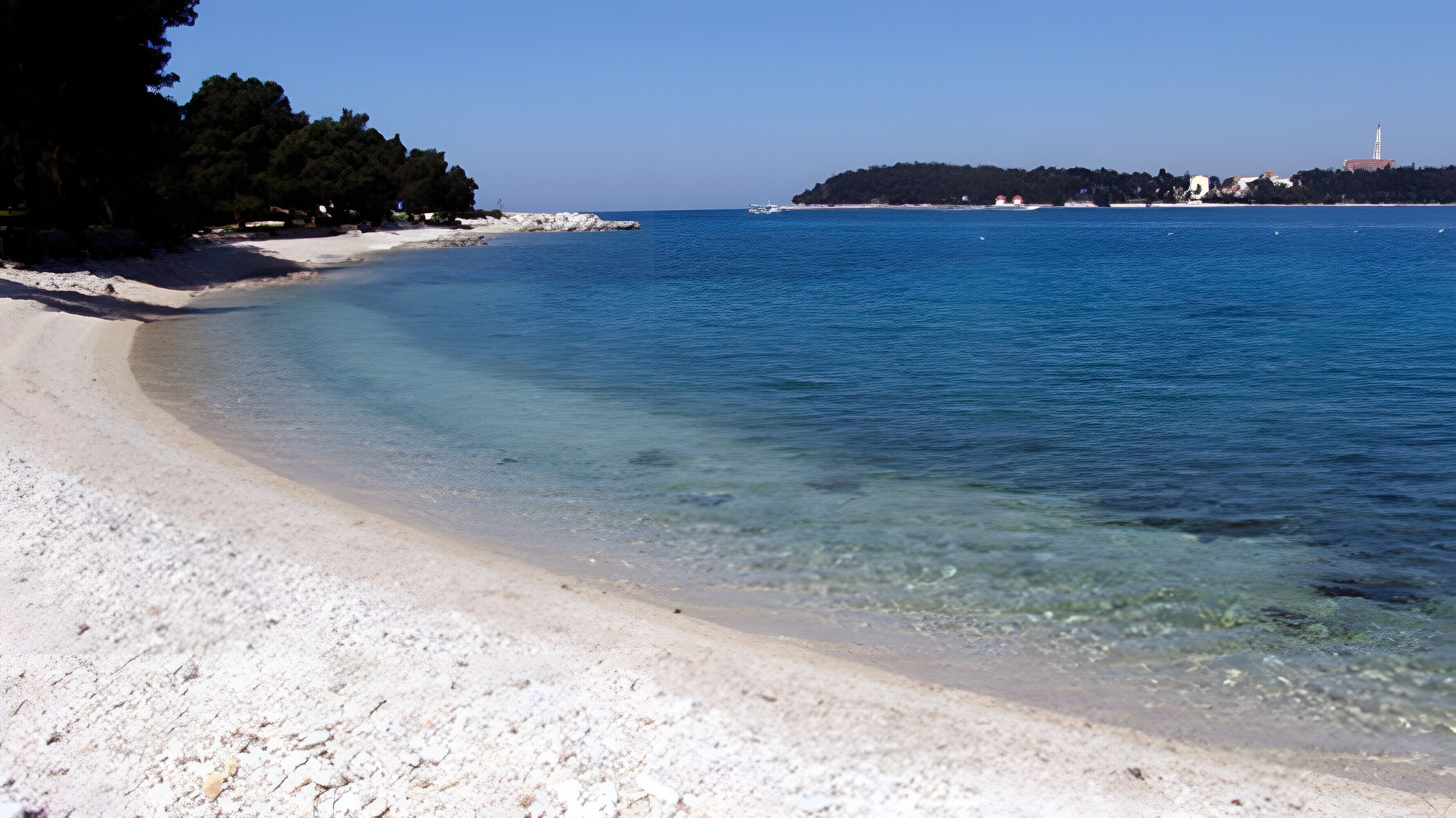 Ein Strand mit klarem Wasser, sandigem Ufer und einer bewaldeten Insel in der Ferne.