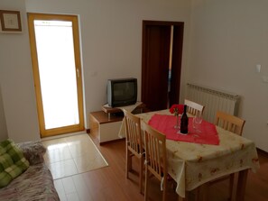 Ein Zimmer mit Esstisch, Stühlen, einem Fernseher auf einem Schrank, einer Couch und einer Heizung.