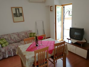A dining area with a table set for two, a sofa, a television, and a window with a view of greenery. - Apartment '2 Pax  On Bike Trails' with Private Terrace, Wi-Fi and Air Conditioning (Rovinj)