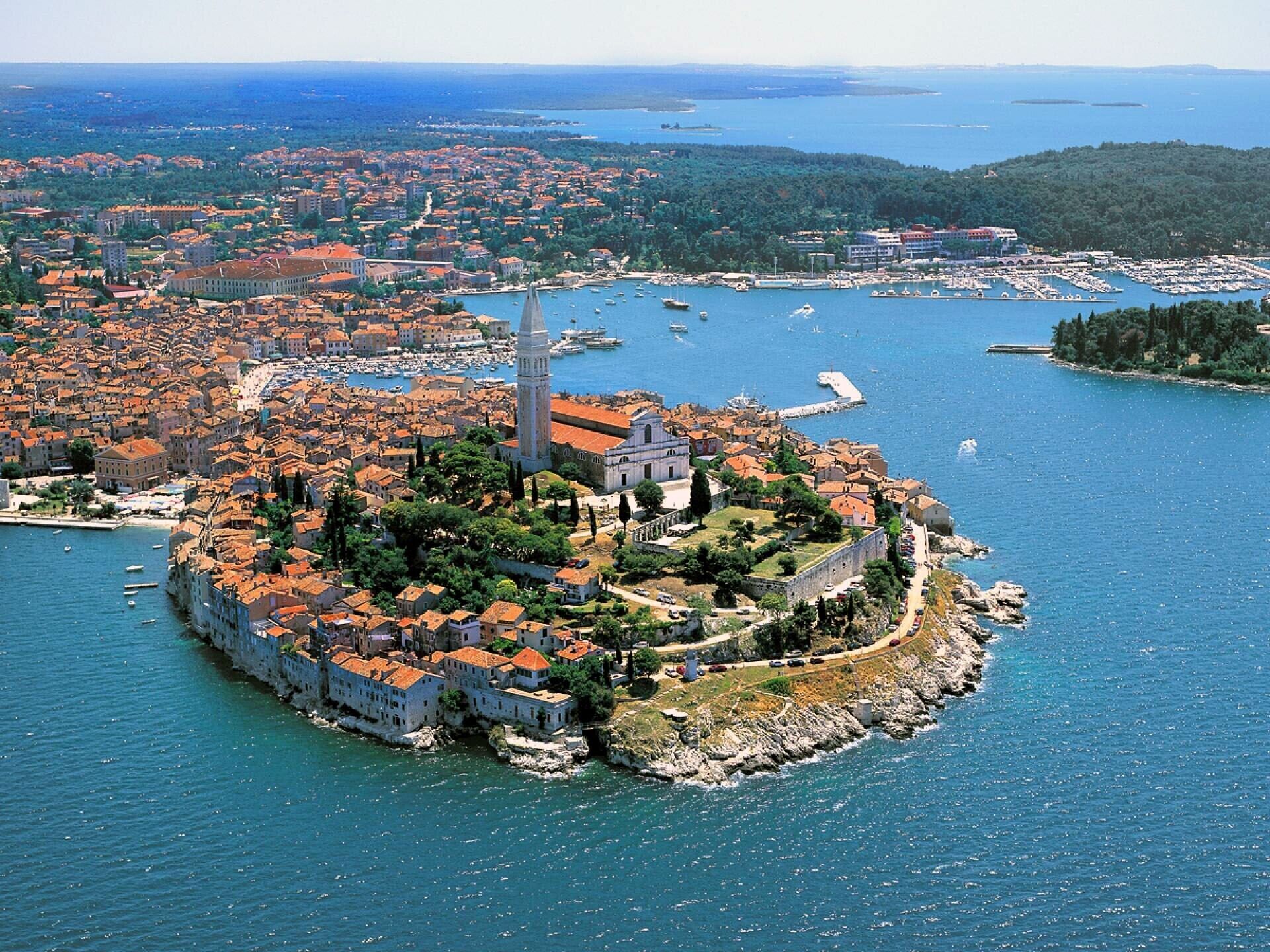 Vue aérienne d’une ville côtière, avec une église et une forteresse imposantes situées sur une petite île, entourées d’eau et d’un quartier urbain densément peuplé.