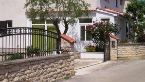A stone wall with a metal gate, a building with a tiled roof, and a tree with green foliage. - Apartment '3 Pax  Near Pebble Beach' with Balcony, Wi-Fi and Air Conditioning (Rovinj)