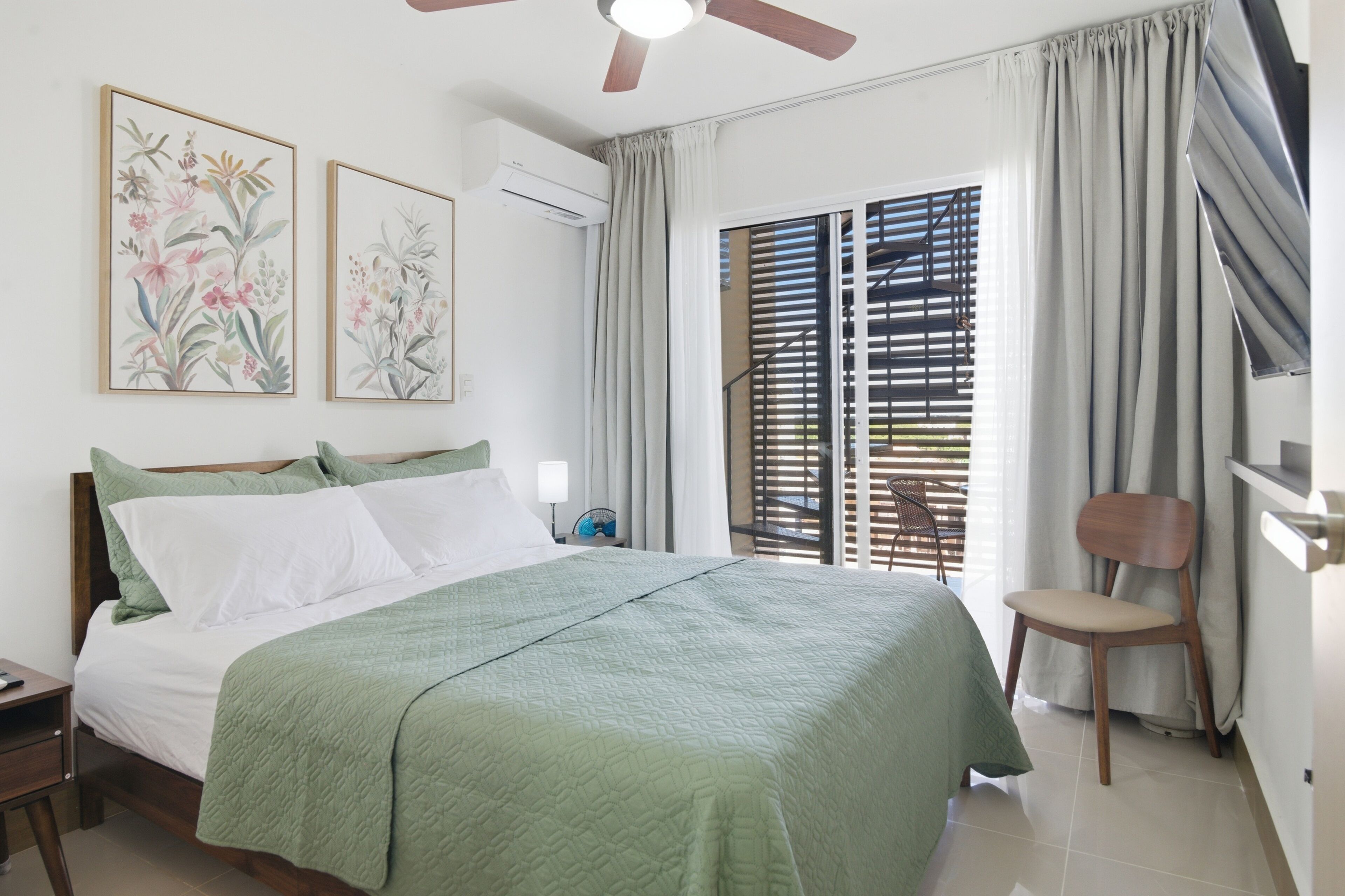 A hotel room with a bed, a chair, a fan, and a window with curtains.