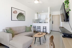 Un salon moderne avec un canapé, une table basse et un téléviseur. Il y a également un coin cuisine équipé d’un réfrigérateur, d’une cuisinière et de quelques tabourets de bar.