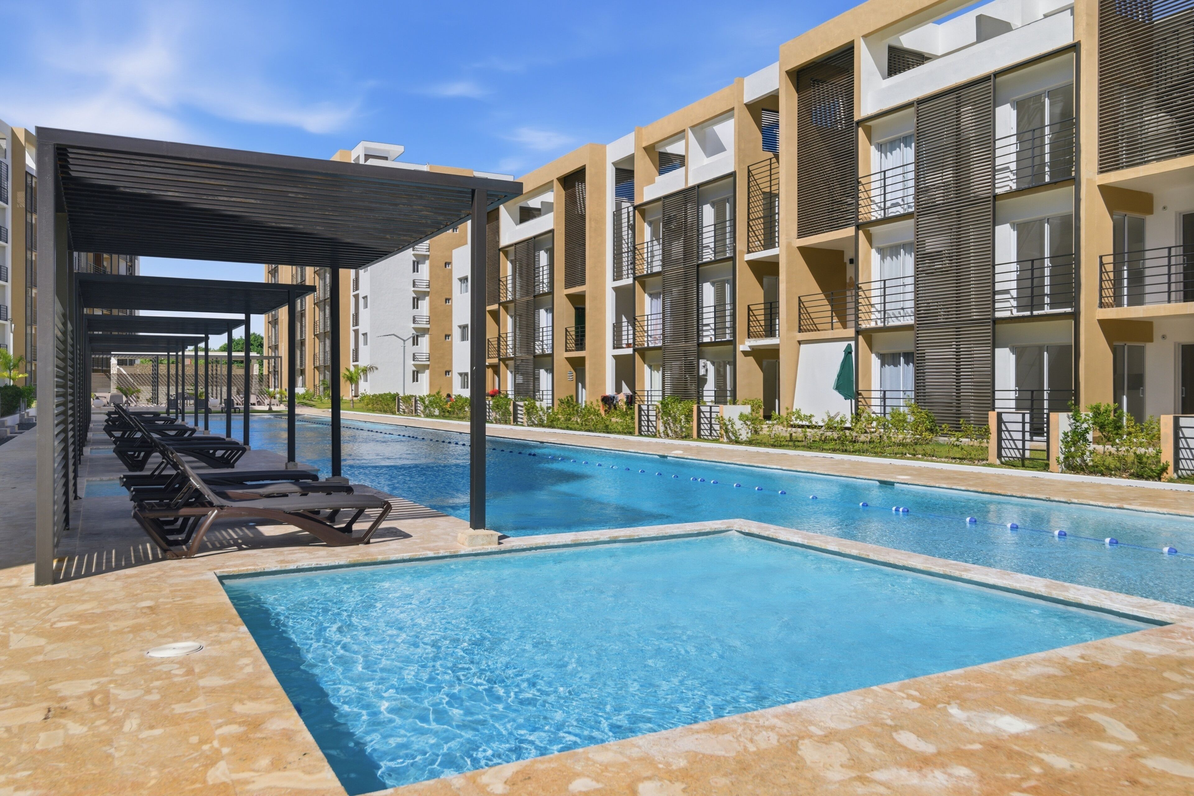 Une piscine dotée de chaises longues et d’un espace de repos couvert, le tout entouré d’immeubles à plusieurs étages avec des balcons.