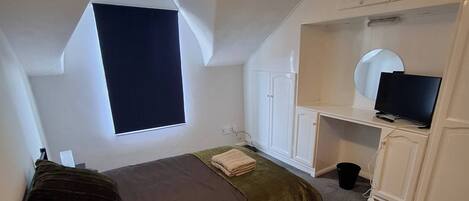 A bedroom with a bed, a desk with a computer, a television, and a window.