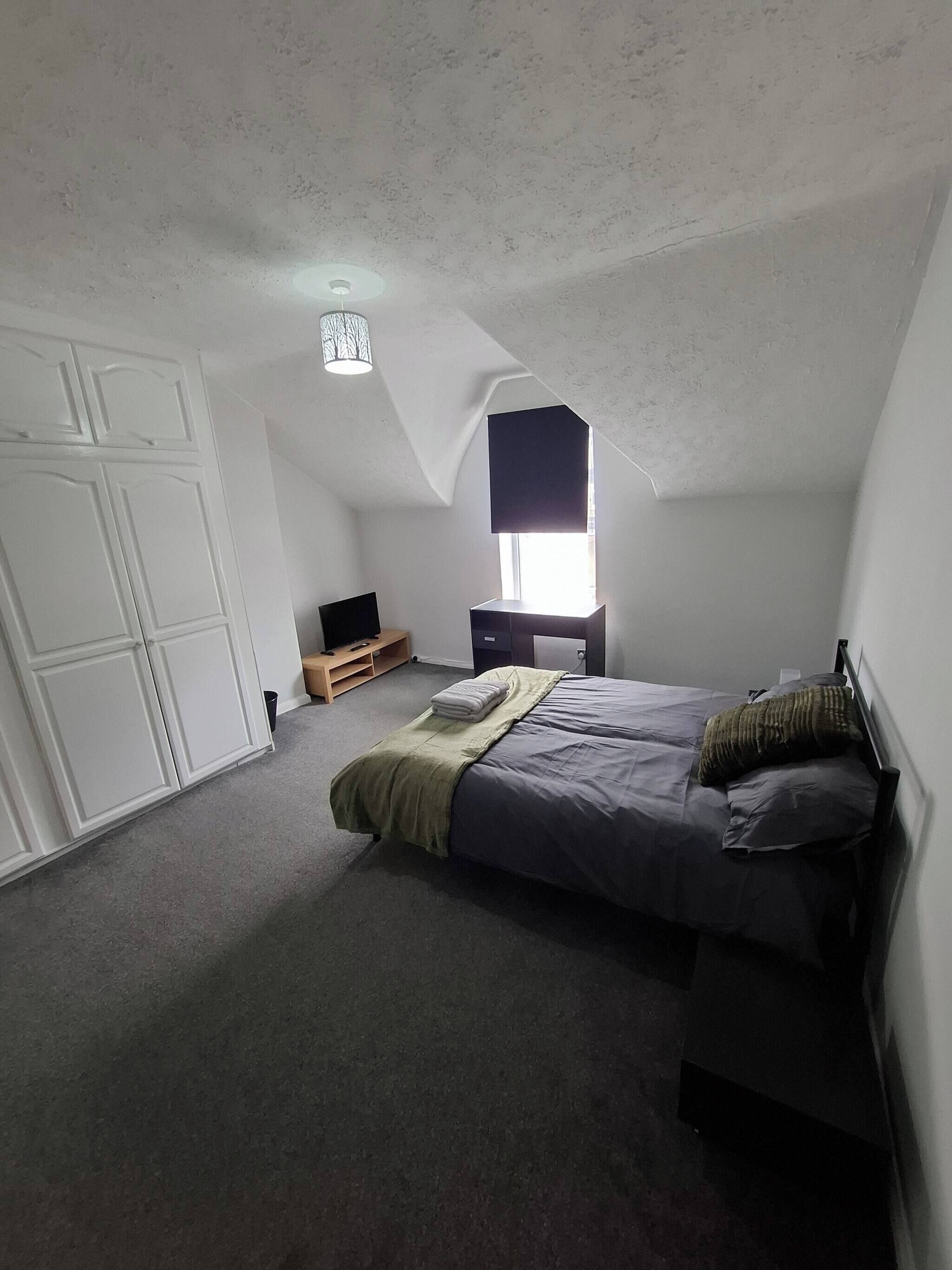 Un dormitorio con techo inclinado, una cama con manta verde, una ventana con persiana negra y un armario blanco.