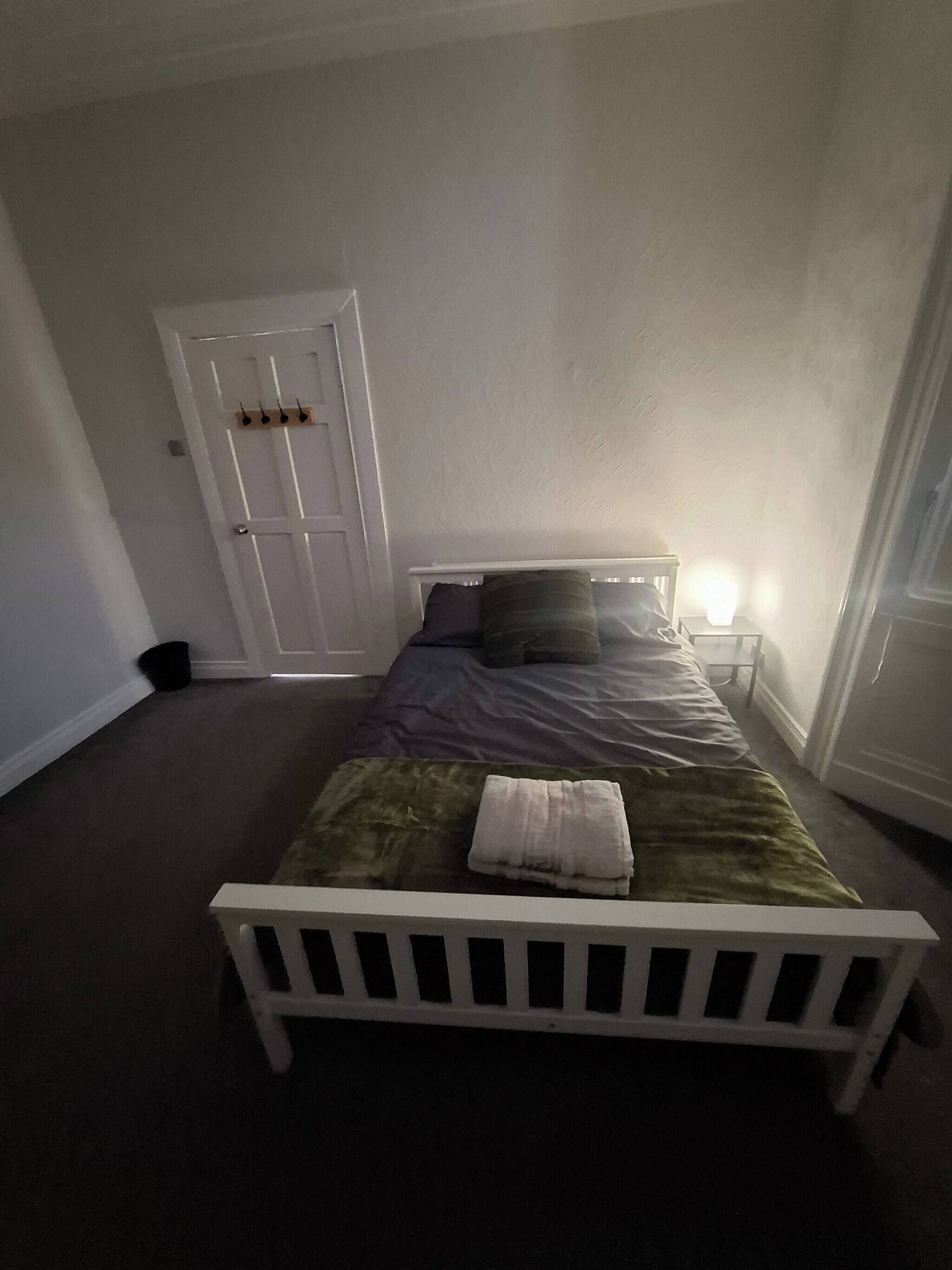 Une chambre avec un lit simple, une tête de lit blanche, une table de chevet avec une lampe et une porte dotée d’un porte-manteau.