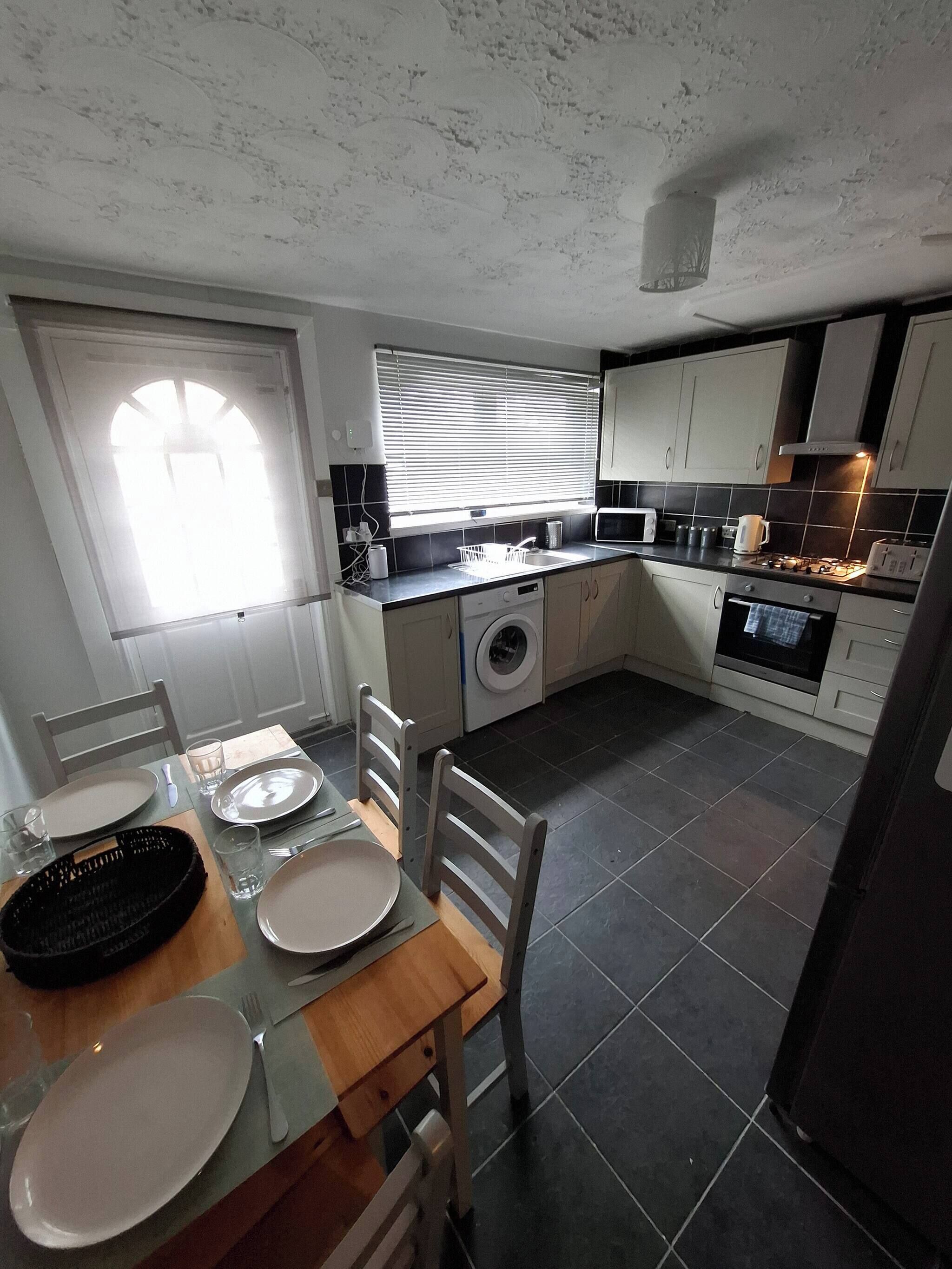 A compact kitchen with a washing machine, a dining area with a table set for two, and a window with blinds.