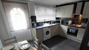 A compact kitchen with a washing machine, a dining area with a table set for two, and a window with blinds.