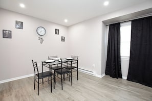 Uma sala de jantar com uma mesa posta para quatro pessoas, um relógio na parede e cortinas pretas na janela.