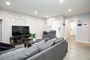 Uma sala de estar moderna com um sofá cinzento, uma televisão de ecrã plano, um console de jogos e uma mesa de centro. Há quadros nas paredes e uma porta que dá para outro quarto.