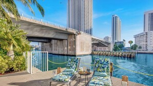 Uima-allasalue aivan veden äärellä, jossa on aurinkotuoleja ja pöytä, silta ja taustalla korkeita rakennuksia.