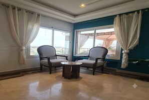 A room with two chairs and a small table, large windows with curtains, and a view of the outdoors.