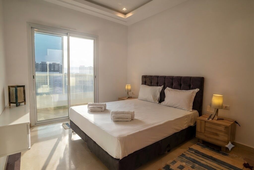 A bedroom with a large bed, two bedside tables, a sliding glass door, and a view of the city.