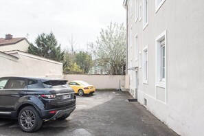 Ein Parkplatz mit zwei Autos, ein Gebäude mit weißen Wänden und Fenstern sowie ein Baum im Hintergrund.
