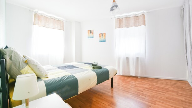 A bedroom with a bed, a bedside table, a lamp, and two windows with curtains. - Evasion Mulhousienne - 2-bedroom apartment with AC, Wi-Fi, and free parking (Mulhouse)