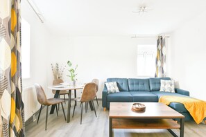 Een moderne woonkamer met een blauwe bank, een houten salontafel en een eetgedeelte met een ronde tafel en stoelen.