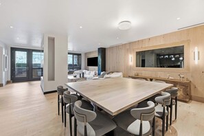 A spacious dining area with a large wooden table and modern chairs, a kitchen with a large window, and a living area with a sofa and a TV. - Solaris 4A East - 4 Bedroom + Den (Vail)