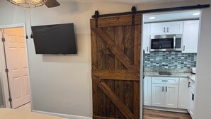 Een slaapkamer met een bed, een aan de muur bevestigde televisie en een schuifdeur van hout die leidt naar een keuken met witte kasten en een magnetron.