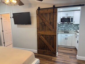 Ein Schlafzimmer mit einem Bett, einem an der Wand befestigten Fernseher und einer Schiebetür vom Typ ‚Barn Door‘, die zu einer Küche mit weißen Schränken und einer Mikrowelle führt.