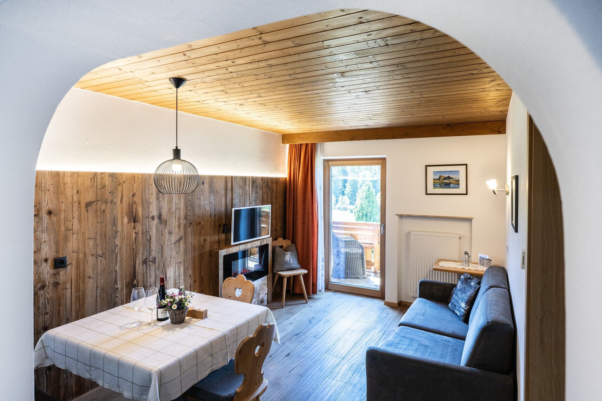 A cozy interior with a wooden ceiling, a dining area with a table and chairs, a sofa, a television, and a window with curtains.