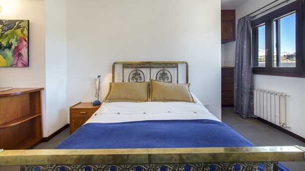 Un dormitorio con cama, mesita de noche, lámpara, un cuadro en la pared y una ventana con cortinas.
