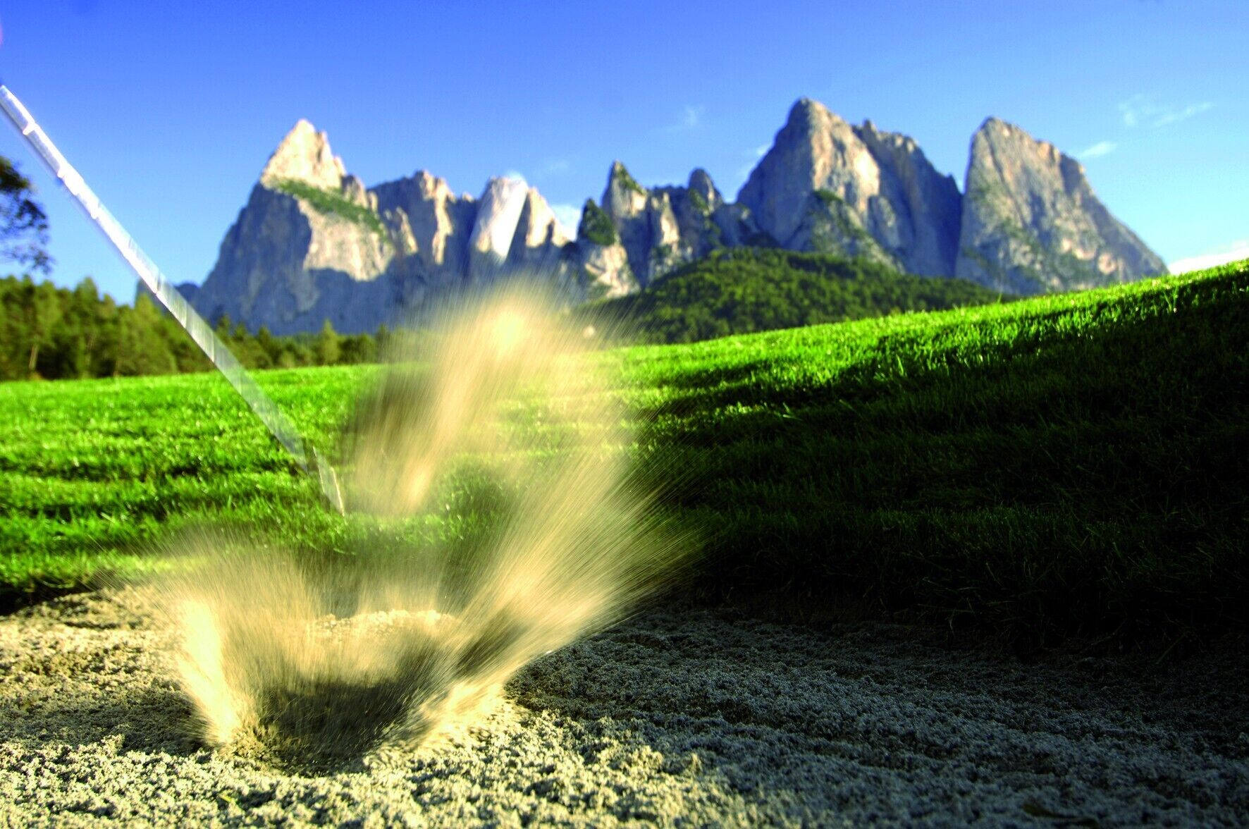 A field with a sprinkler in motion, a clear blue sky, and a mountain range in the background.