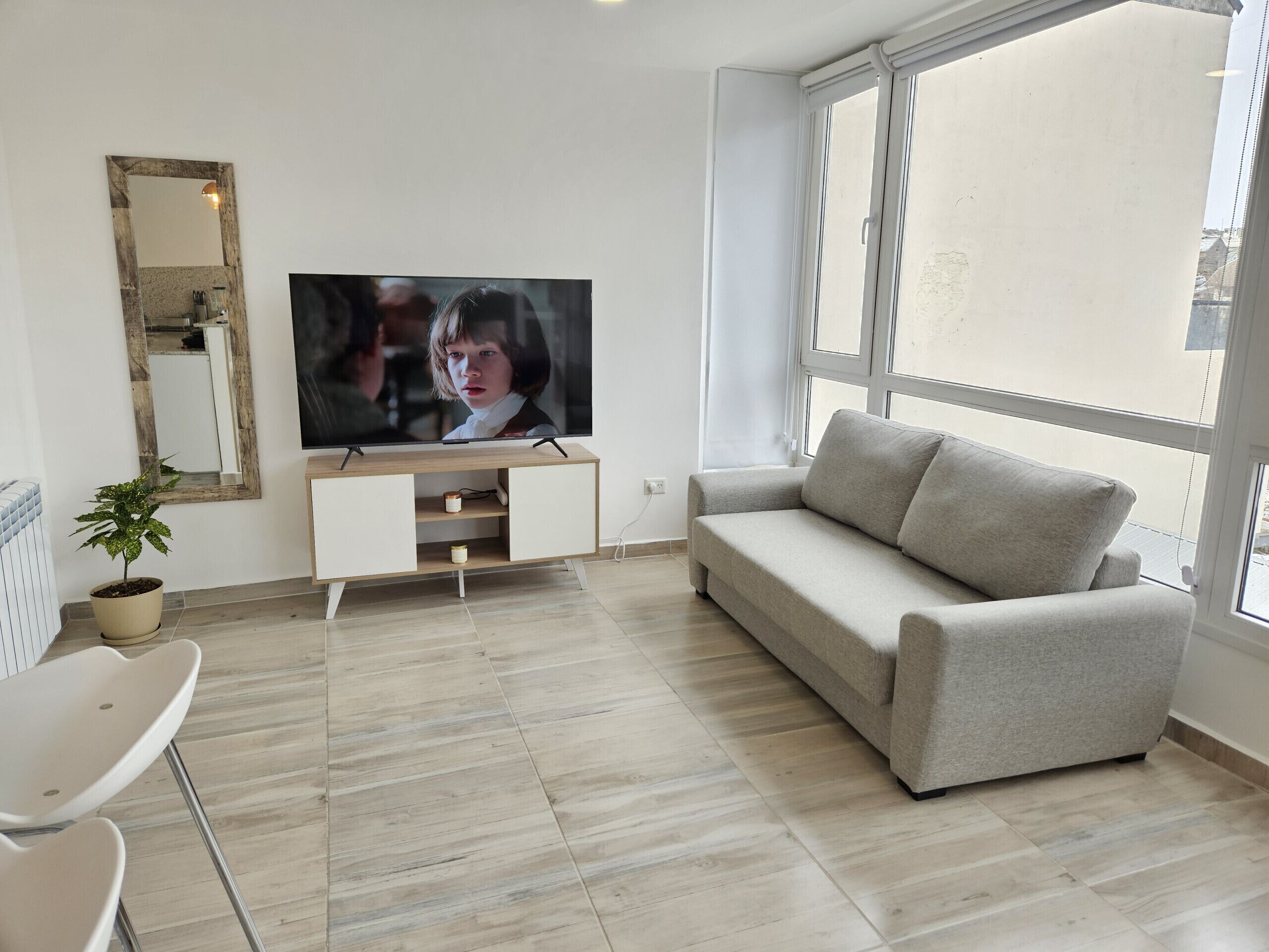Una sala de estar con un televisor de pantalla plana, un sofá, una silla, una planta en maceta y una ventana.