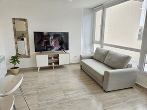 Ein Wohnzimmer mit Flachbildfernseher, einer Couch, einem Sessel, einer Zimmerpflanze und einem Fenster.
