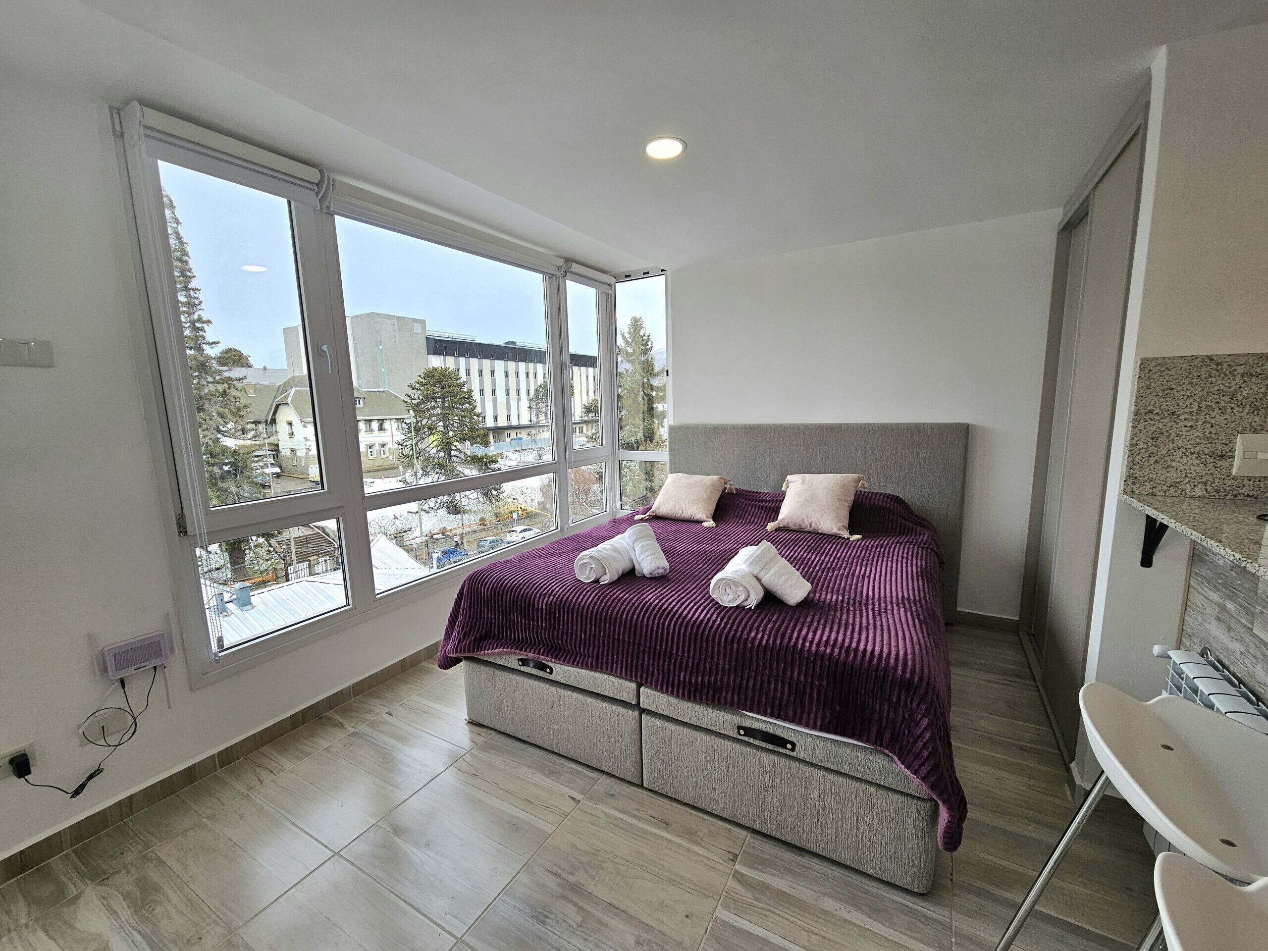 Un dormitorio con cama, ventana grande y vista a edificios.
