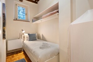 A small, compact bedroom with a single bed, a bedside table with a lamp, a radiator, a window with frosted glass, and a shelf with towels. - Apartment "Casa Conrad - Lucca" with Wi-Fi and Air Conditioning (Lucca)