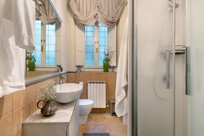 Ein Badezimmer mit einem großen weißen Waschbecken, einer Toilette, einer Duschkabine und einem Fenster mit Vorhängen.