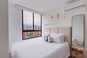Ein Schlafzimmer mit großem Fenster, einem Bett mit weißer Bettwäsche, einem Nachttisch, einem Spiegel und einer wandmontierten Klimaanlage.