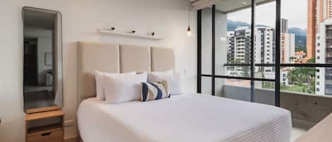 A modern hotel room with a large bed, a bedside table, a mirror, and a view of the cityscape through large windows.
